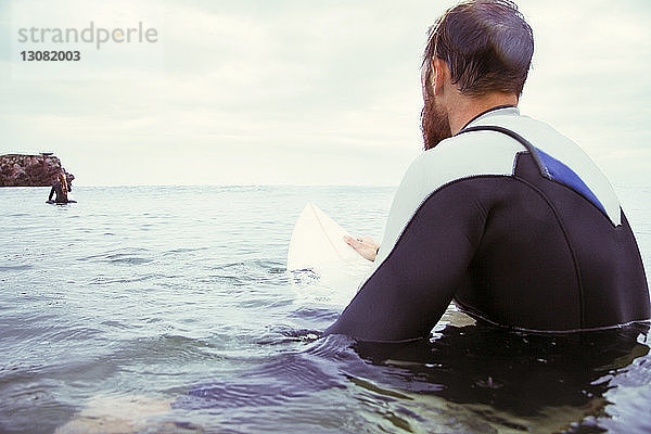 Rückansicht eines männlichen Surfers  der auf einem Surfbrett im Meer vor bewölktem Himmel sitzt
