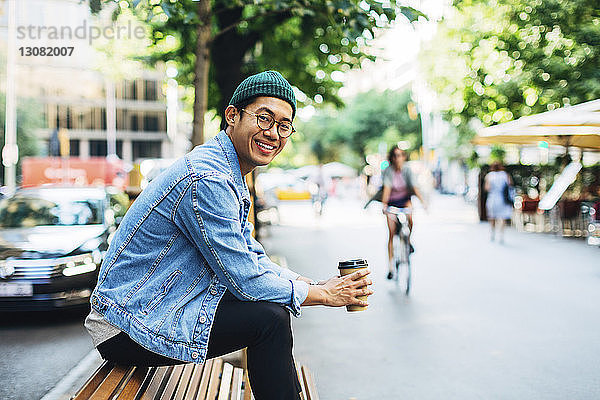 Porträt eines glücklichen Mannes  der einen Einwegbecher hält  während er auf einer Bank auf dem Bürgersteig sitzt