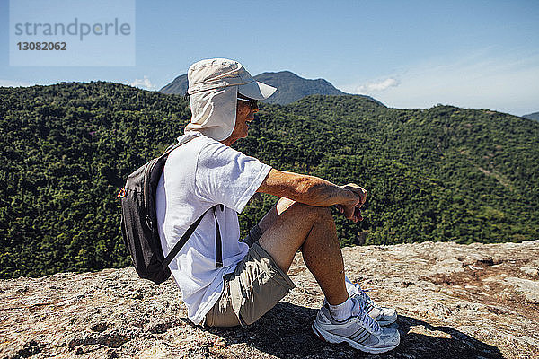 Seitenansicht eines männlichen Wanderers  der am sonnigen Tag auf einem Berg vor blauem Himmel sitzt