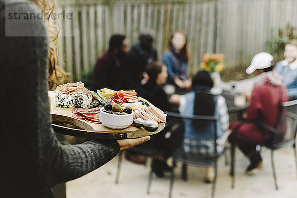 Frau trägt Essen auf Holzbrettchen  während sich Freunde im Garten vergnügen