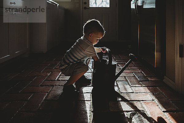 Seitenansicht eines kleinen Jungen  der eine Gießkanne hält  während er zu Hause auf dem Boden kauert