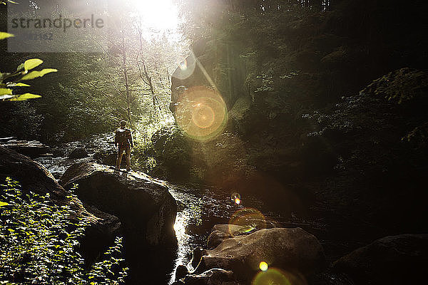 Wanderer in voller Länge auf einem Felsen am Bach im Wald an einem sonnigen Tag stehend
