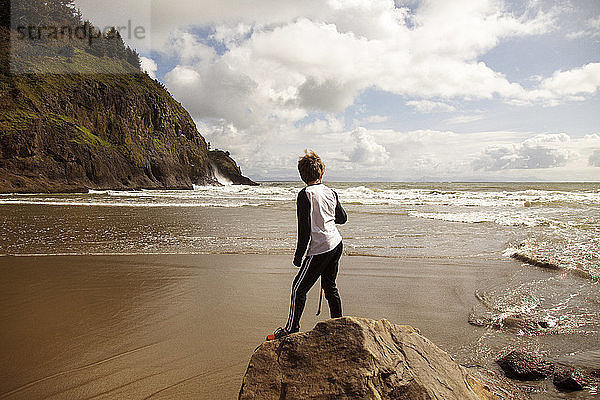 Rückansicht eines Jungen  der auf einem Felsen am Meeresufer vor bewölktem Himmel steht