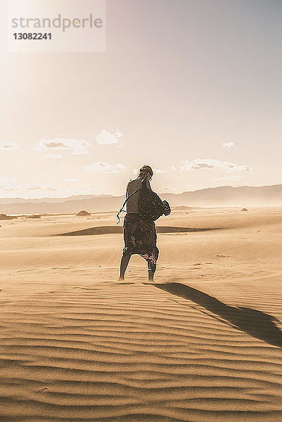 Rückansicht einer Frau mit Rucksack  die bei Sonnenuntergang auf Sand am Strand gegen den Himmel läuft