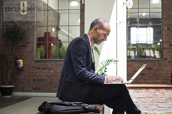 Seitenansicht eines Geschäftsmannes  der einen Laptop-Computer benutzt  während er auf einem Sitz gegen sein Büro sitzt