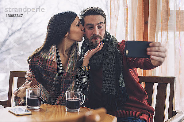 Frau küsst  während ihr Freund sich bei Tisch im Cafe mit sich selbst beschäftigt
