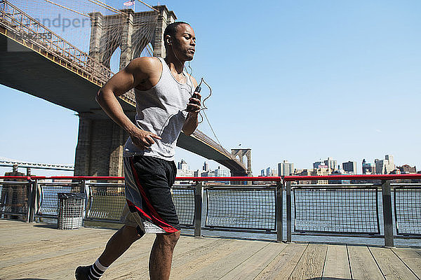 Mann hört Musik und joggt auf der Strandpromenade gegen die Brooklyn Bridge
