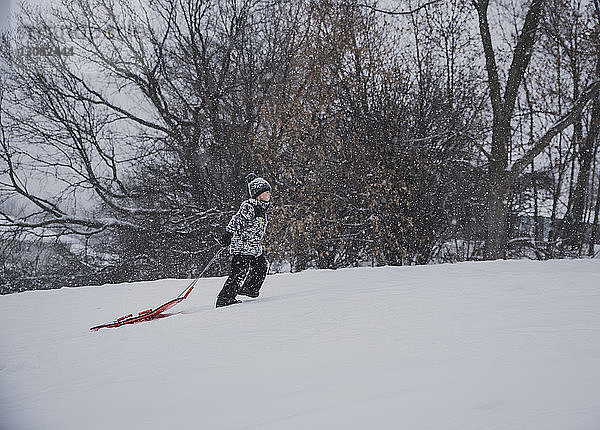 Seitenansicht eines Jungen  der einen Schlitten zieht  während er auf einem verschneiten Feld gegen kahle Bäume läuft