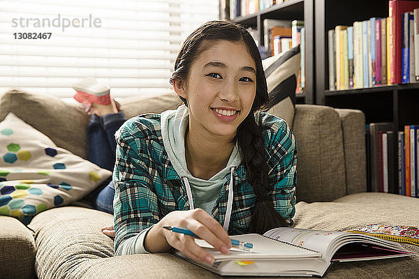 Porträt einer lächelnden Teenagerin  die Hausaufgaben macht  während sie zu Hause auf der Couch liegt