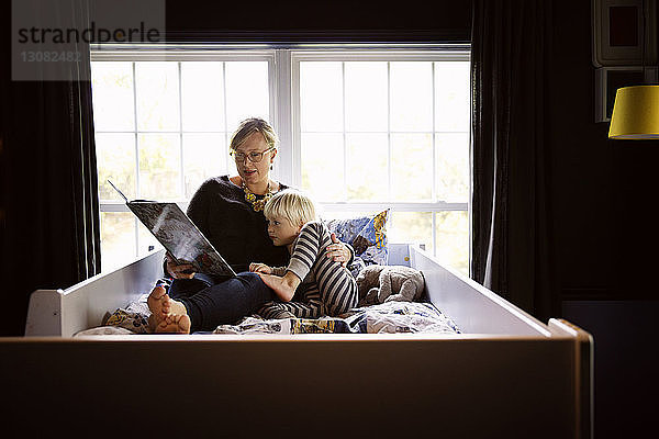 Mutter liest dem neugierigen Sohn ein Buch vor  während er zu Hause im Bett am Fenster sitzt