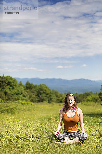 Frau meditiert  während sie auf dem Feld vor bewölktem Himmel sitzt