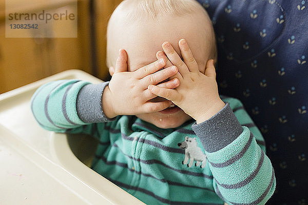 Verspielter kleiner Junge  der sich die Augen zuhat  während er zu Hause auf einem Hochstuhl Kuckuck spielt