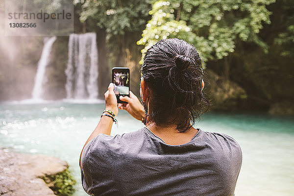 Rückansicht eines Mannes  der mit einem Smartphone einen Wasserfall fotografiert