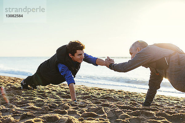 Seitenansicht von Vater und Sohn beim Liegestützen  während sie bei Sonnenuntergang am Strand gegen den klaren Himmel Händchen halten