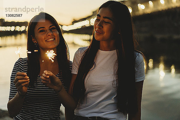 Glückliche Freunde verbrennen Wunderkerzen  während sie bei Sonnenuntergang am Fluss Arno gegen den Himmel sitzen