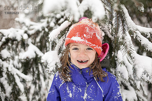 Porträt eines fröhlichen Mädchens in warmer Kleidung  das vor einem schneebedeckten Baum steht