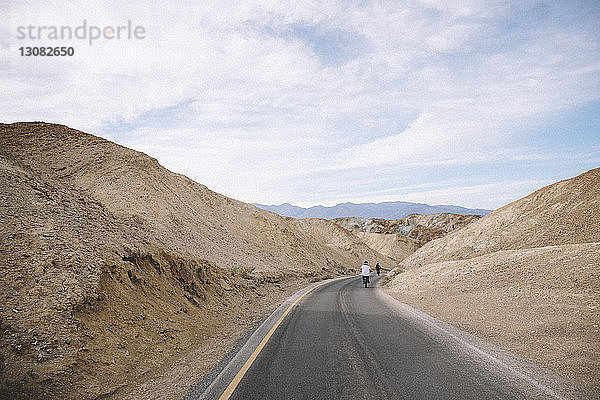 Rückansicht von Vater und Sohn beim Fahrradfahren auf einer Wüstenstraße vor bewölktem Himmel