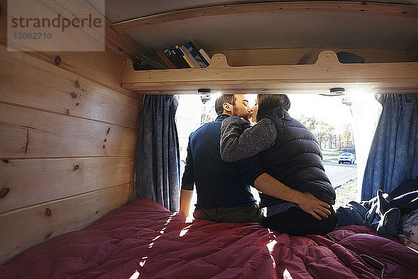 Rückansicht eines Pärchens  das sich küsst  während es im Wohnmobil auf dem Bett sitzt