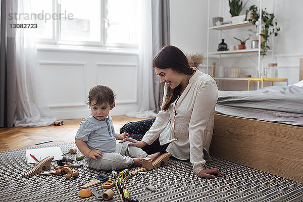 Glückliche Mutter schaut dem kleinen Jungen zu  der im Schlafzimmer mit Spielzeug auf dem Teppich spielt