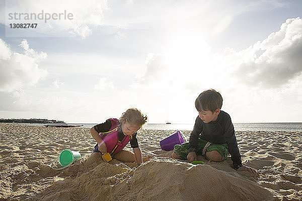 Geschwister spielen mit Sand am Strand gegen den Himmel