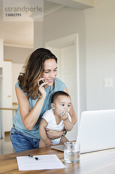 Mutter telefoniert  während sie ein Baby trägt Junge schaut auf Laptop-Computer