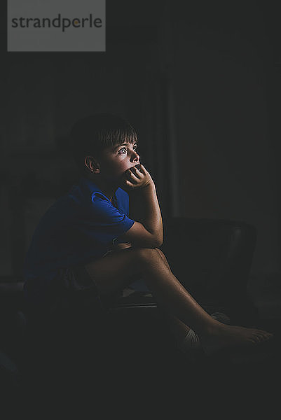 Seitenansicht eines nachdenklichen Jungen mit Hand am Kinn  der auf einem Stuhl in der Dunkelkammer sitzt