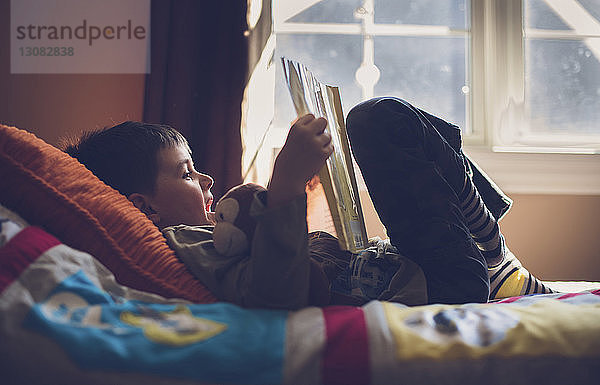 Junge mit ausgestopftem Spielzeug-Lesebuch  während er zu Hause auf dem Bett liegt
