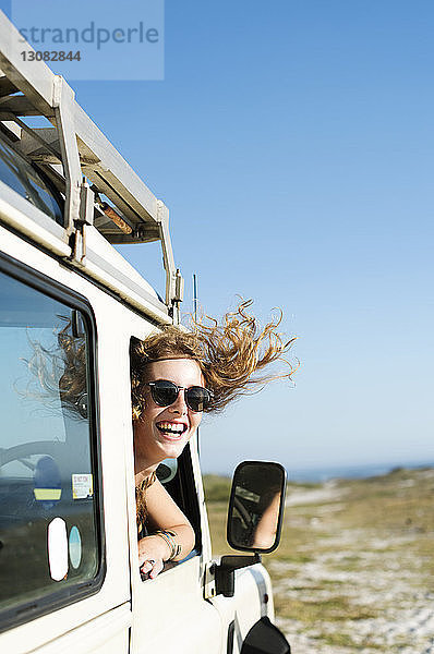 Glückliche Frau mit Sonnenbrille  die am sonnigen Tag durch das Autofenster schaut