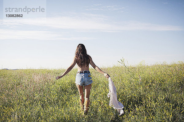 Rückansicht einer Frau  die ein Hemd hält und auf einem Grasfeld gegen den Himmel läuft