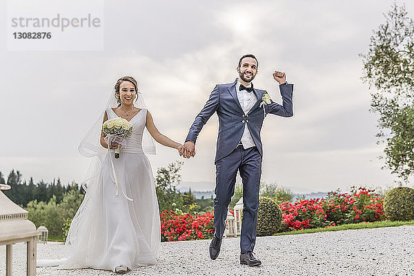Glückliches frisch verheiratetes Paar hält sich an den Händen  während es auf einem Fußweg gegen den bewölkten Himmel geht