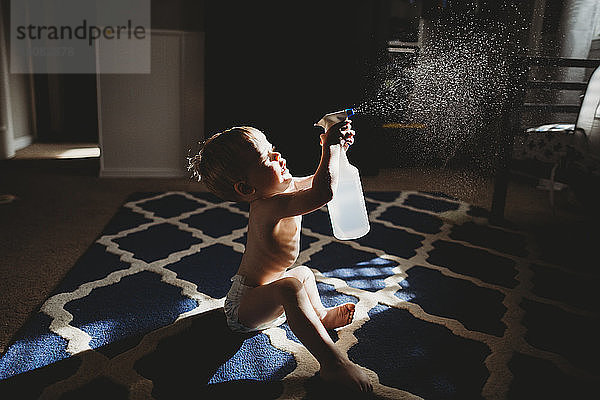 Seitenansicht eines Jungen ohne Hemd  der Wasser versprüht  während er zu Hause auf dem Teppich sitzt