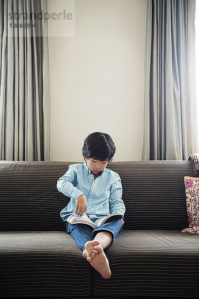 Junge liest Buch  während er zu Hause auf dem Sofa an der Wand sitzt