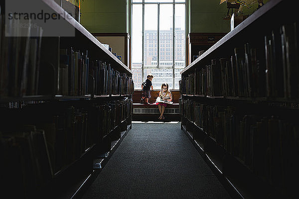 Junge sieht Schwester an  die in der Bibliothek am Fenster liest