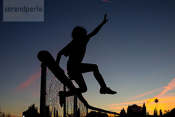 Silhouette eines sportlichen Mädchens  das in der Abenddämmerung gegen den Himmel über den Zaun springt