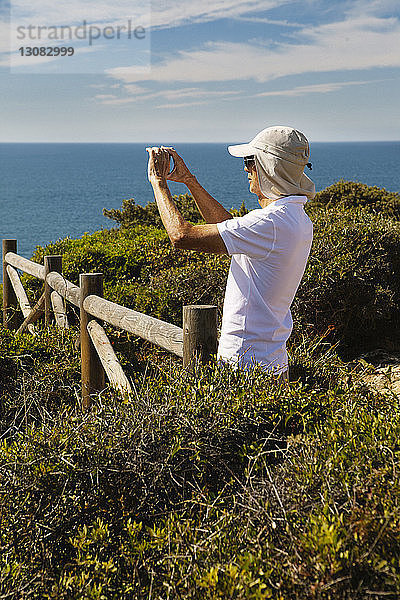 Mann fotografiert Meer durch Telefon  während er auf einer Klippe gegen den Himmel steht