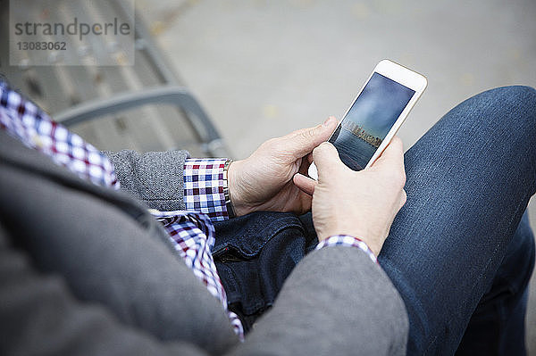 Mittelsektion eines Mannes  der ein Mobiltelefon benutzt  während er im Park auf einer Bank sitzt