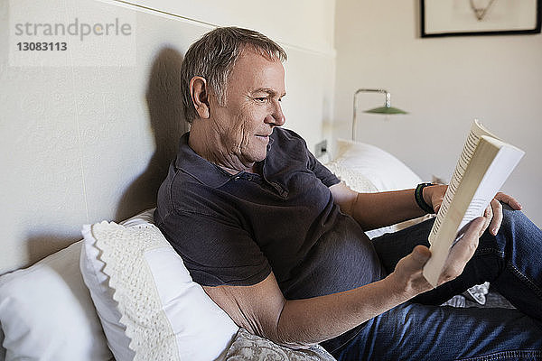 Älterer Mann liest zu Hause auf dem Bett sitzend ein Buch