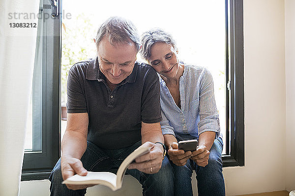 Glückliche reife Frau  die ein Smartphone benutzt  während der Mann zu Hause sitzt und ein Buch liest