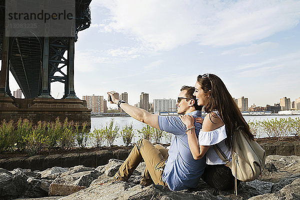 Glückliches Paar beim Selbermachen  während es auf Felsen am East River gegen den Himmel sitzt