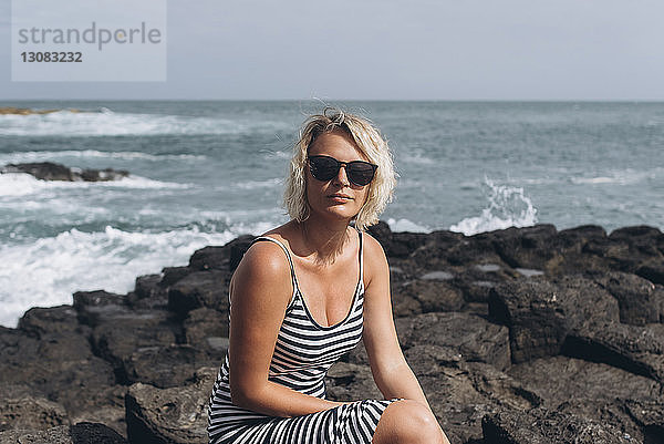 Porträt einer selbstbewussten Frau mit Sonnenbrille  die auf Felsen am Strand vor dem Meer sitzt