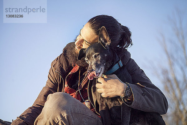 Anhänglicher Mann umarmt Hund  während er vor klarem blauen Himmel sitzt