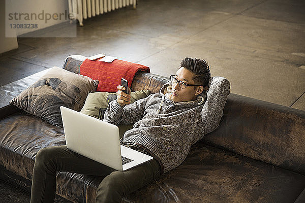 Hochwinkelansicht eines Geschäftsmannes mit Laptop-Computer  der das Telefon benutzt  während er im Kreativbüro auf dem Sofa liegt