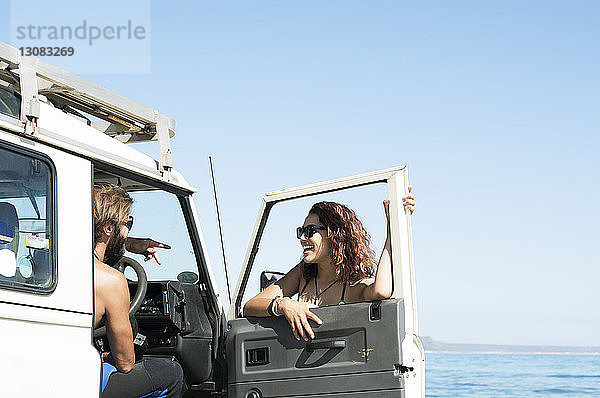 Frau unterhält sich mit einem männlichen Freund  der an einem sonnigen Tag im Geländewagen am Strand sitzt