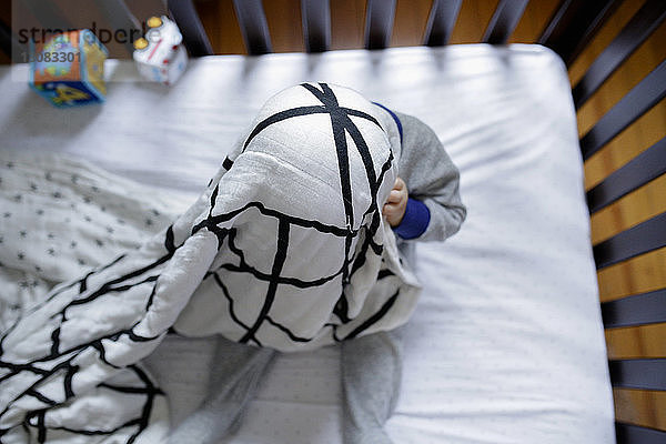 Draufsicht auf einen verspielten kleinen Jungen  der sich in einer Decke versteckt  während er zu Hause in der Krippe Kuckuck spielt