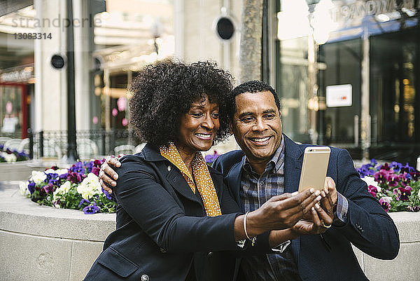 Lächelndes Paar  das sich über ein Smartphone selbstständig macht  während es in der Stadt auf einem Sitzplatz sitzt