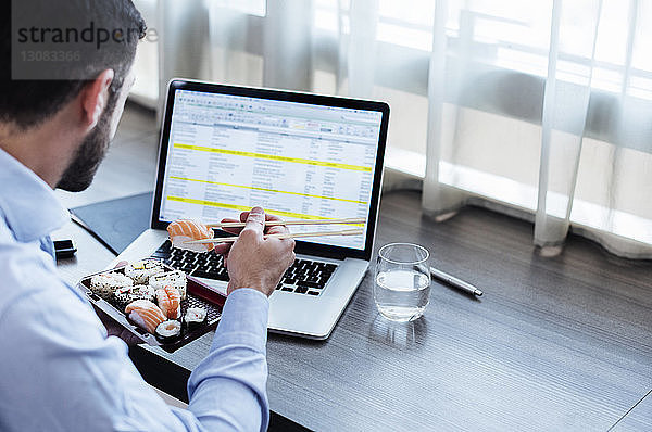Seitenansicht eines Geschäftsmannes  der Sushi isst  während er seinen Laptop auf dem Fensterbrett eines Hotelzimmers benutzt