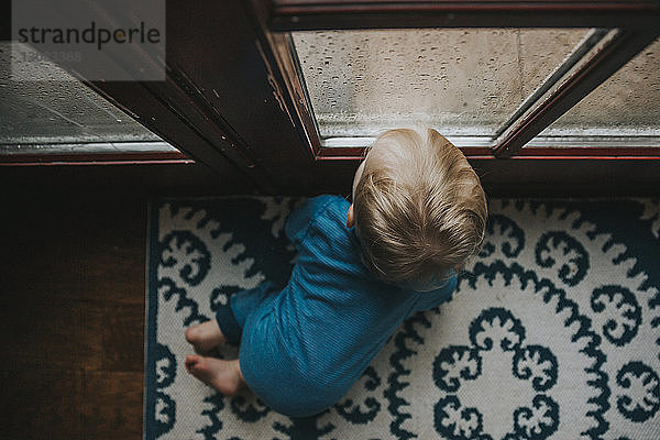 Hochwinkelansicht eines kleinen Jungen  der durch ein nasses Fenster zu Hause schaut