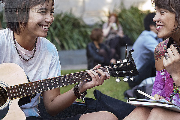 Lächelnde Frau hört einem Freund zu  der im Park Gitarre spielt