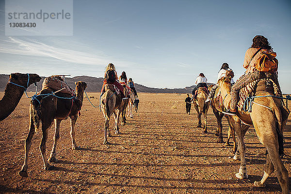 Rückansicht von Touristen  die bei Sonnenschein auf Kamelen in der Wüste gegen den Himmel reiten