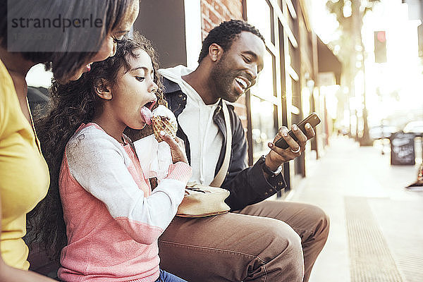 Mädchen leckt Eiscreme  während die Eltern am Bürgersteig sitzen